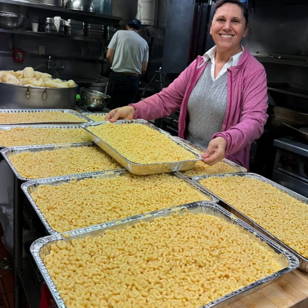 A smiling woman in a commercial kitchen holds a large tray of macaroni and cheese.