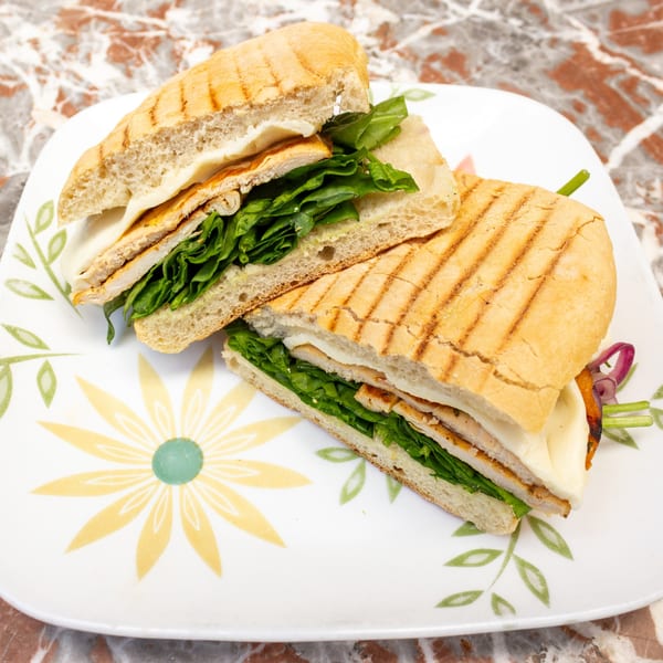 Grilled chicken panini with melted cheese and fresh spinach on a white plate with a floral design.