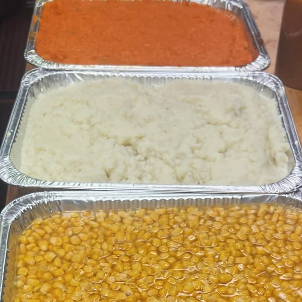 Three foil trays of food: orange mashed sweet potatoes, white mashed potatoes, and yellow corn.