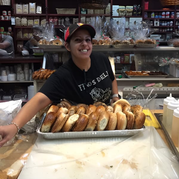 A smiling Tino's Delicatessen employee in a cap stands behind a tray of bagels.